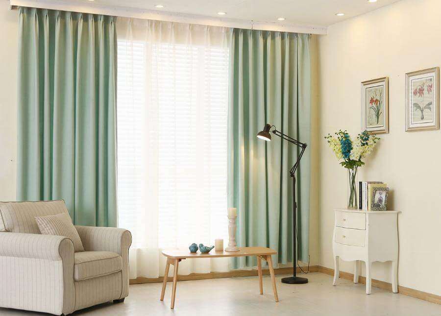 Sage green curtains on a ceiling track parted over a white sheer, with a striped armchair and floor lamp